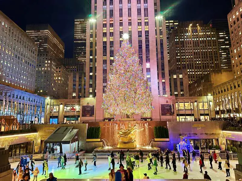 ice skating rink with gold statue behind and behind that large tree coovered in colroed lights and behind that tall art deco style skyscraper